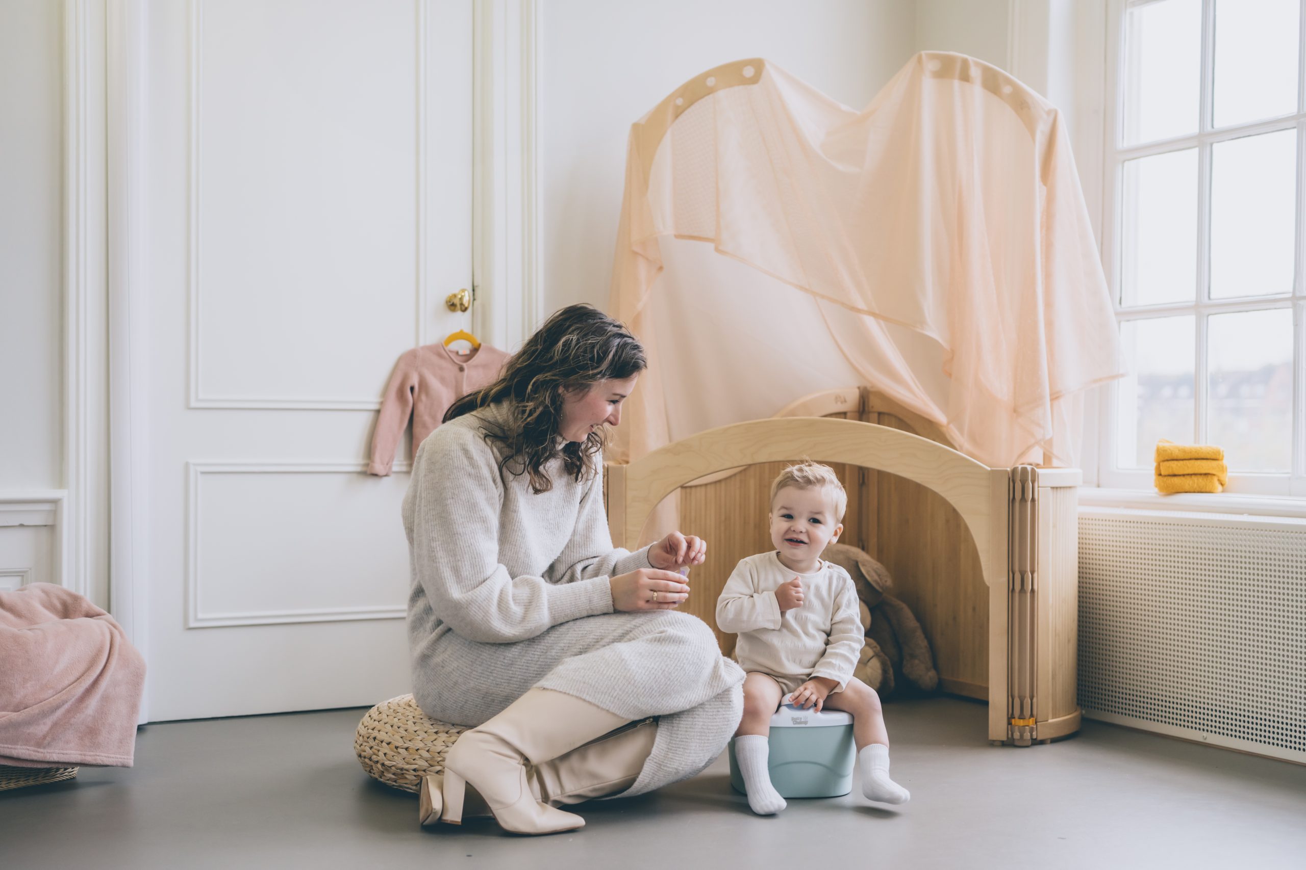 A young child, around 2 years old, sitting on a light blue plastic potty seat/potty chair with their pants pulled down, smiling and looking happy. The child's mother is sitting on a chair next to the potty for potty training toddler/toilet training, looking at the child and also smiling because it is the right potty training age for child. Both the mother and child appear happy and relaxed. There are potty training book, toys and clothes visible in the background. They use potty training app Go Potty which is a free potty training app