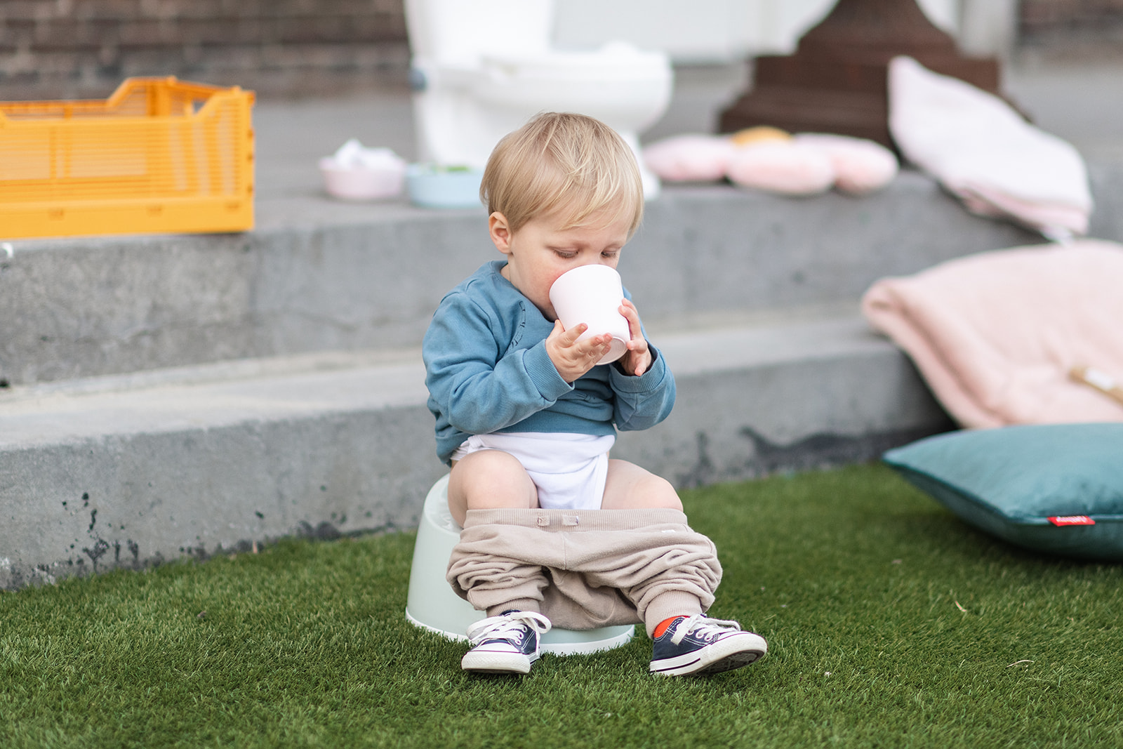 A young child, around 2 years old, sitting on a white plastic potty with their pants pulled down. The child is holding a plastic cup with both hands and is taking a sip of water. There are potty training book and toys visible in the background. They use potty training app Go Potty which is a free potty training app