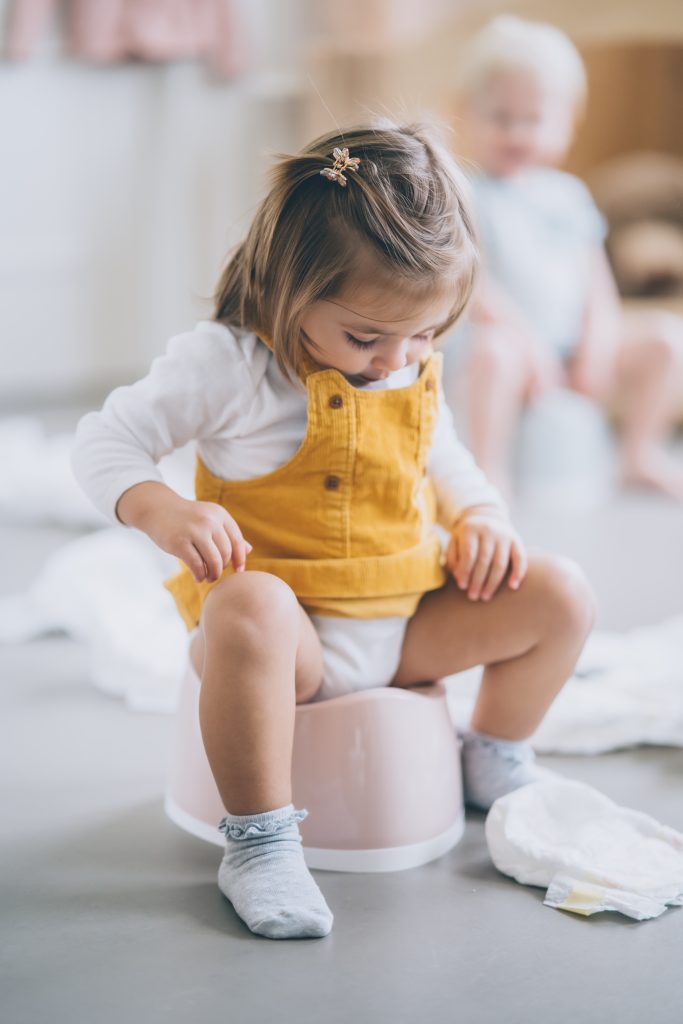 A little girl sits on a potty while looking down at her dress. She appears curious and focused, with her hands in her lap. The potty is pink colored. She is sitting on the potty for poop/pee/wee/poo during potty training.