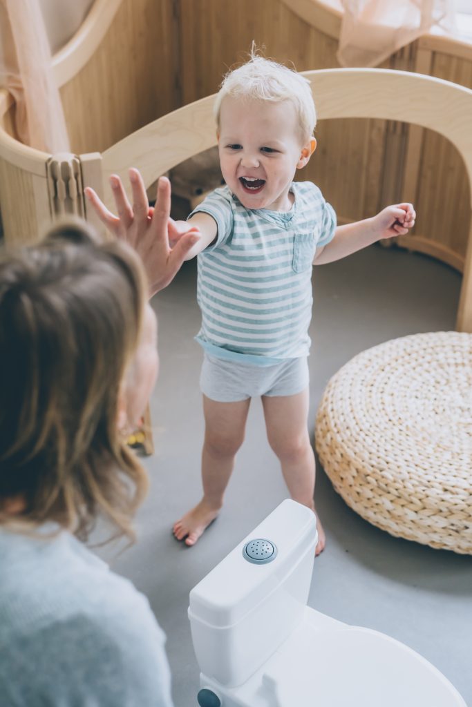 A small child, approximately 2 years old, standing besides a white plastic potty with his pants pulled up, looking happy and excited. The child's mother is sitting beside the child and holding out her hand for a high five. The mother and child are both smiling. The baby is excitedly doing the high five. 