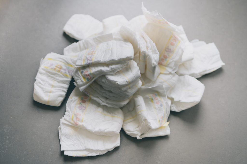 A stack of white disposable diapers/nappies. The diapers/nappies are of similar size. The background is a solid color, which appears to be light gray.