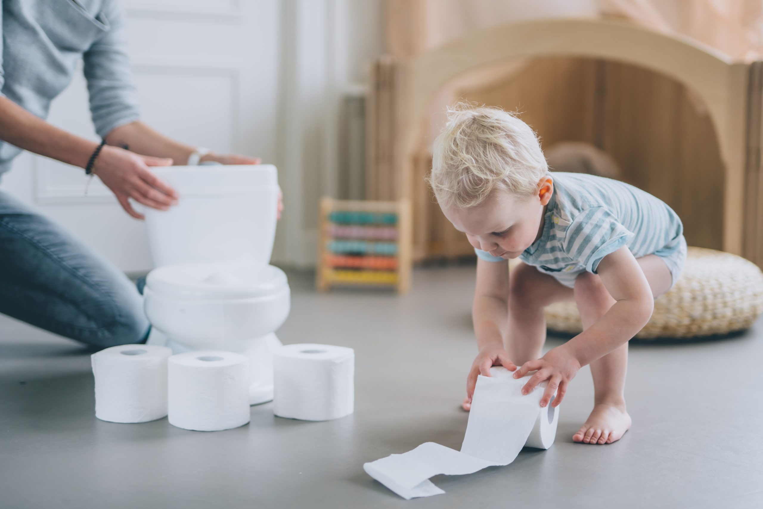 baby is lowering down to pick up a tissue paper roll during potty training. a toilet seat/ potty chair can be seen in the background with tissues and wipes used for poop, pee, wee, or poo.