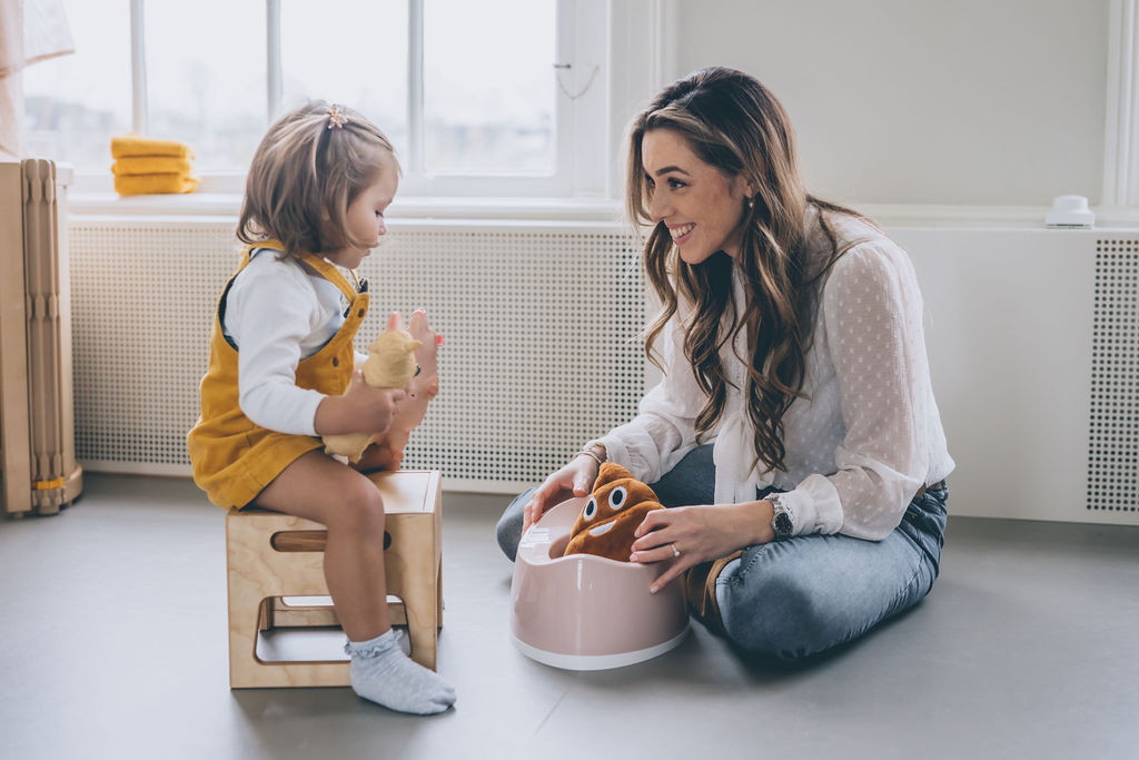 child sitting on a pink potty during potty training. Tissue/wipes and toys can be seen in the background. child and mother playing during potty training / toilet training. both are happy because child doesn't refuse or child won't sit on potty child is happy