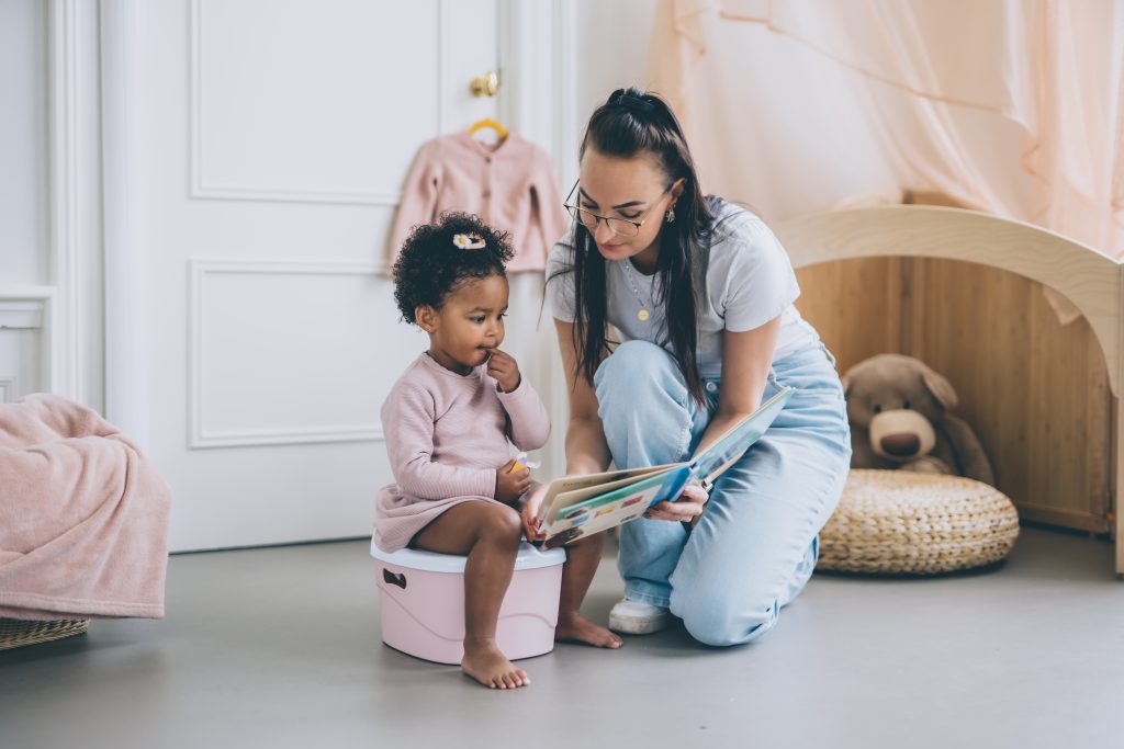 child sitting on a pink potty during potty training. Tissue/wipes and toys can be seen in the background. child and mother playing during potty training / toilet training. both are happy because child doesn't refuse or child won't sit on potty child is happy