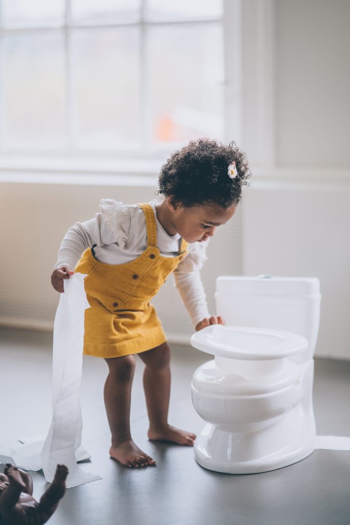 A child playfully exploring a toilet seat, lifting it up and down with curiosity. The child's face is filled with interest and exploration, showing their fascination with the toilet. The toilet seat is clean and closed, and the child is safely engaged in their exploration. She is trying to overcome her potty training fears