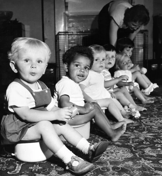 Black and whote picture of six children potty training by sitting in a row on potties in a nursery in 1955.